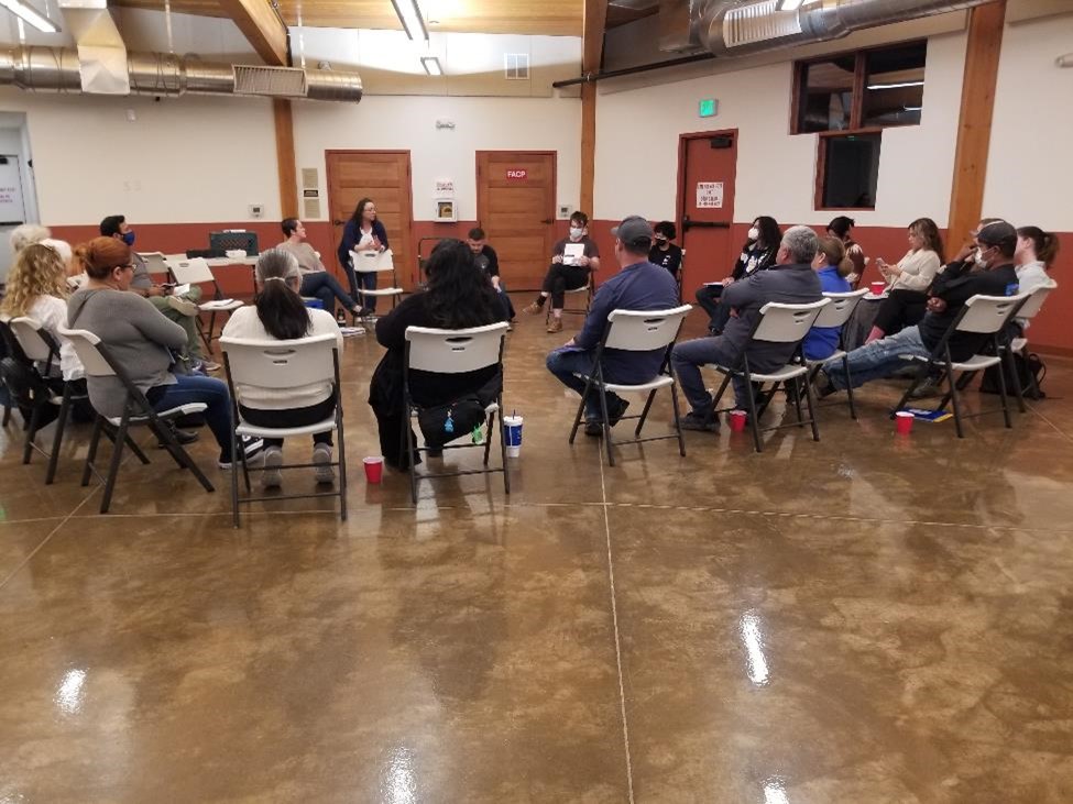 Around 25 people sitting in chairs in a circle listening to a legal presentation. Their backs are facing the camera.